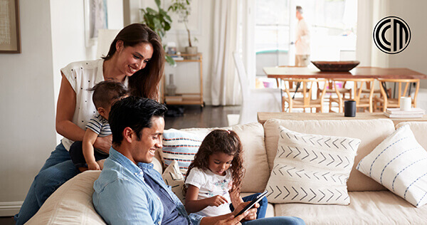 A family sitting together on a couch reads news about the CCM Community Promise program expansion on a tablet.